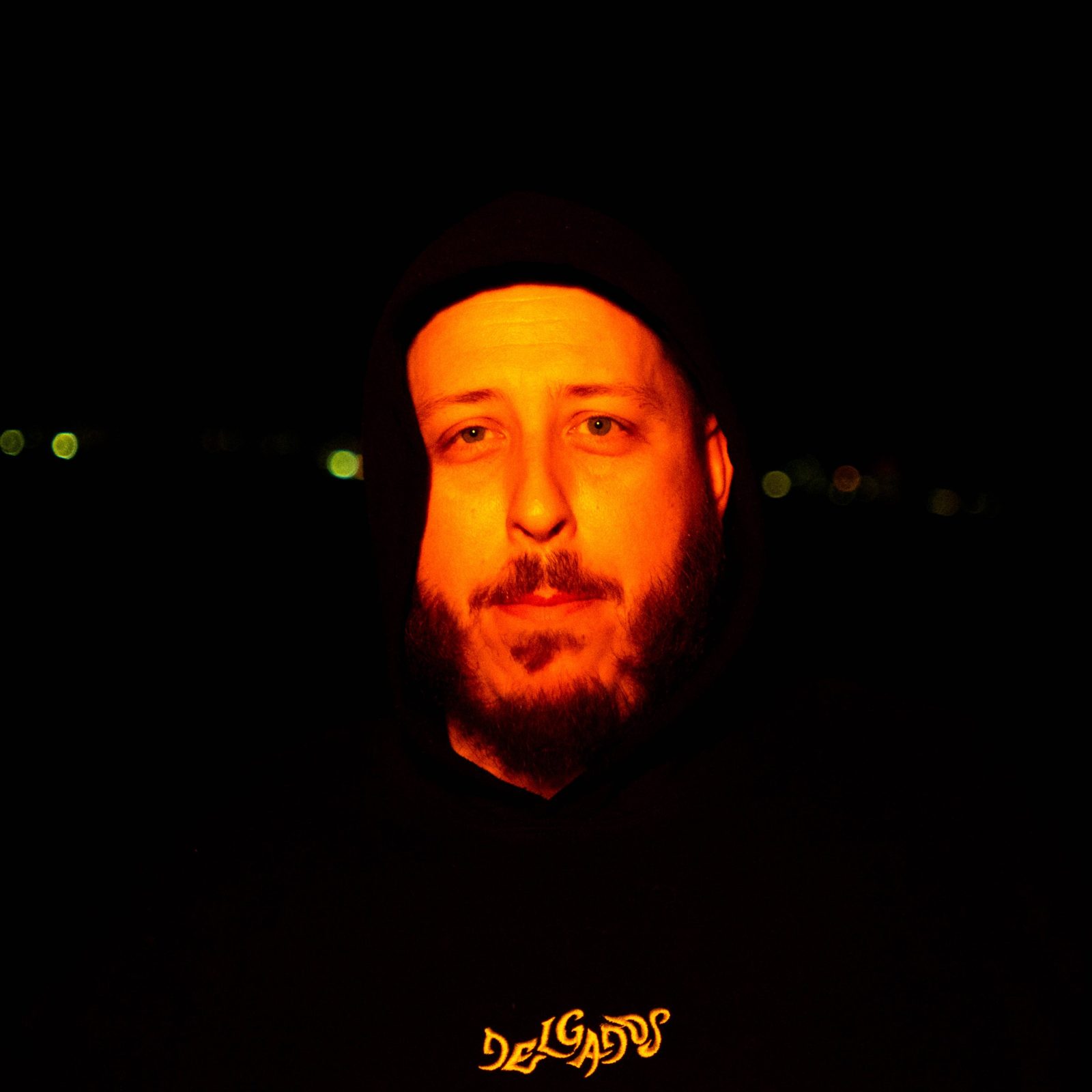 Fenix at night in a black hood, his face lit orange, city lights behind him